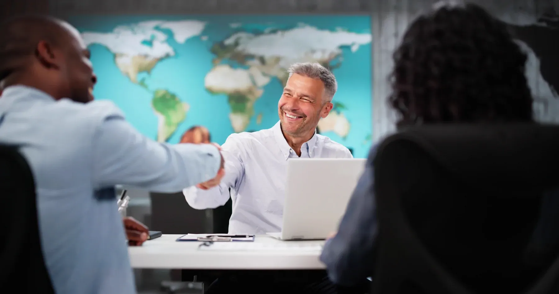 Colleagues shaking hands across a table in a meeting room, a world map on the wall behind them.