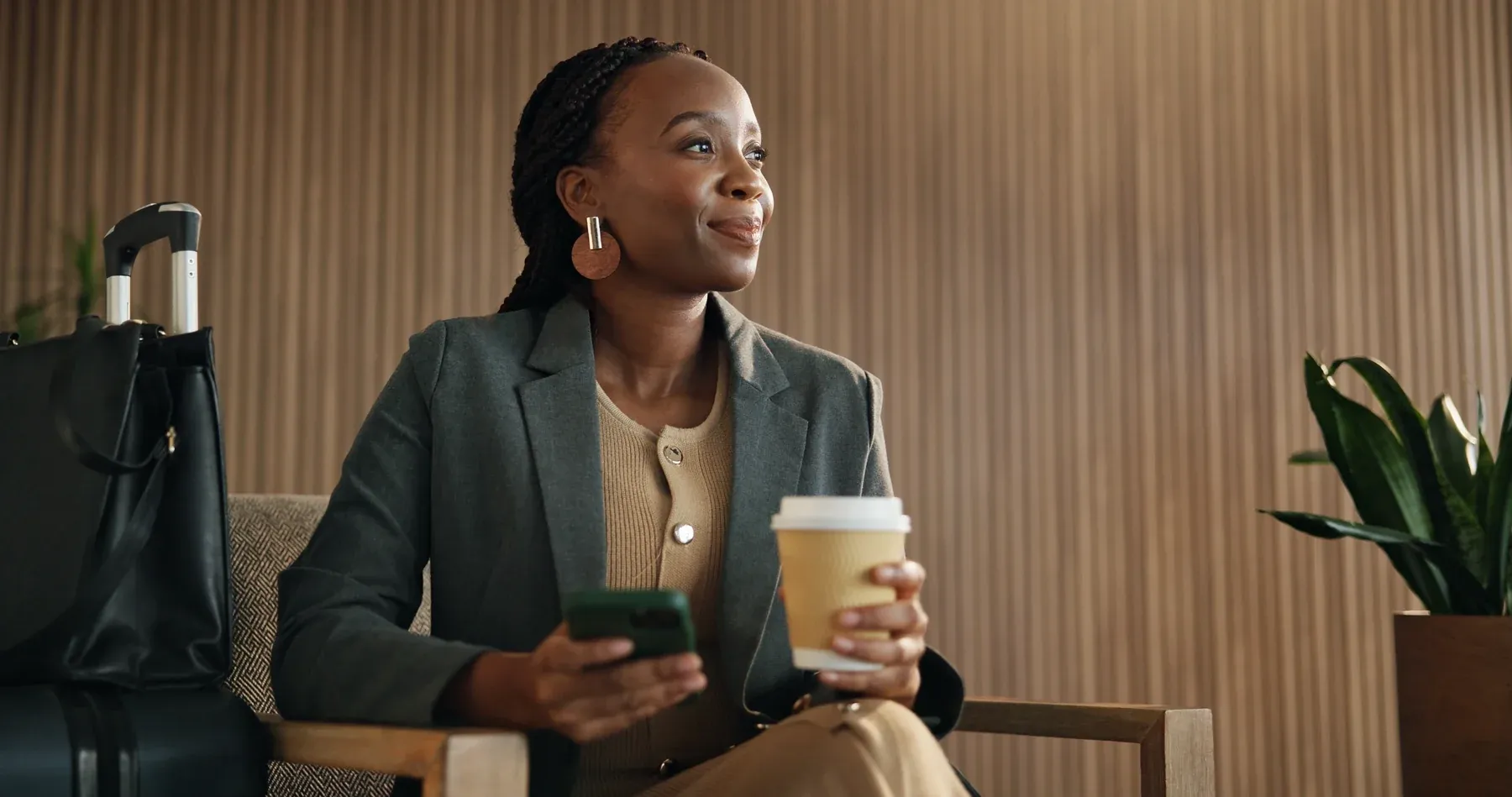 A business traveller seated in a lounge with her luggage, checking her phone over a coffee.